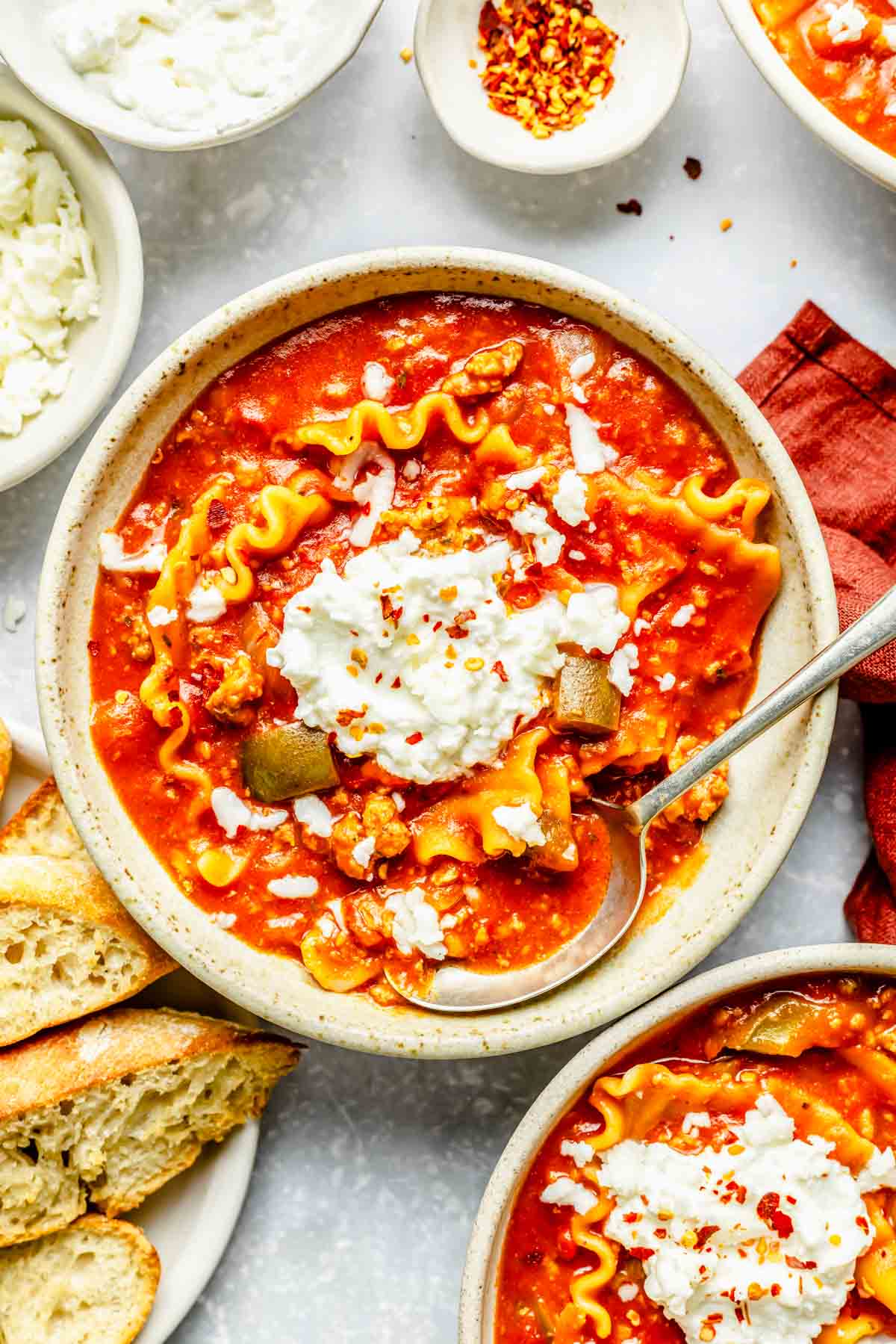 a bowl of lasagna soup with a dollop of ricotta cheese on top and a metal spoon next to toasted bread