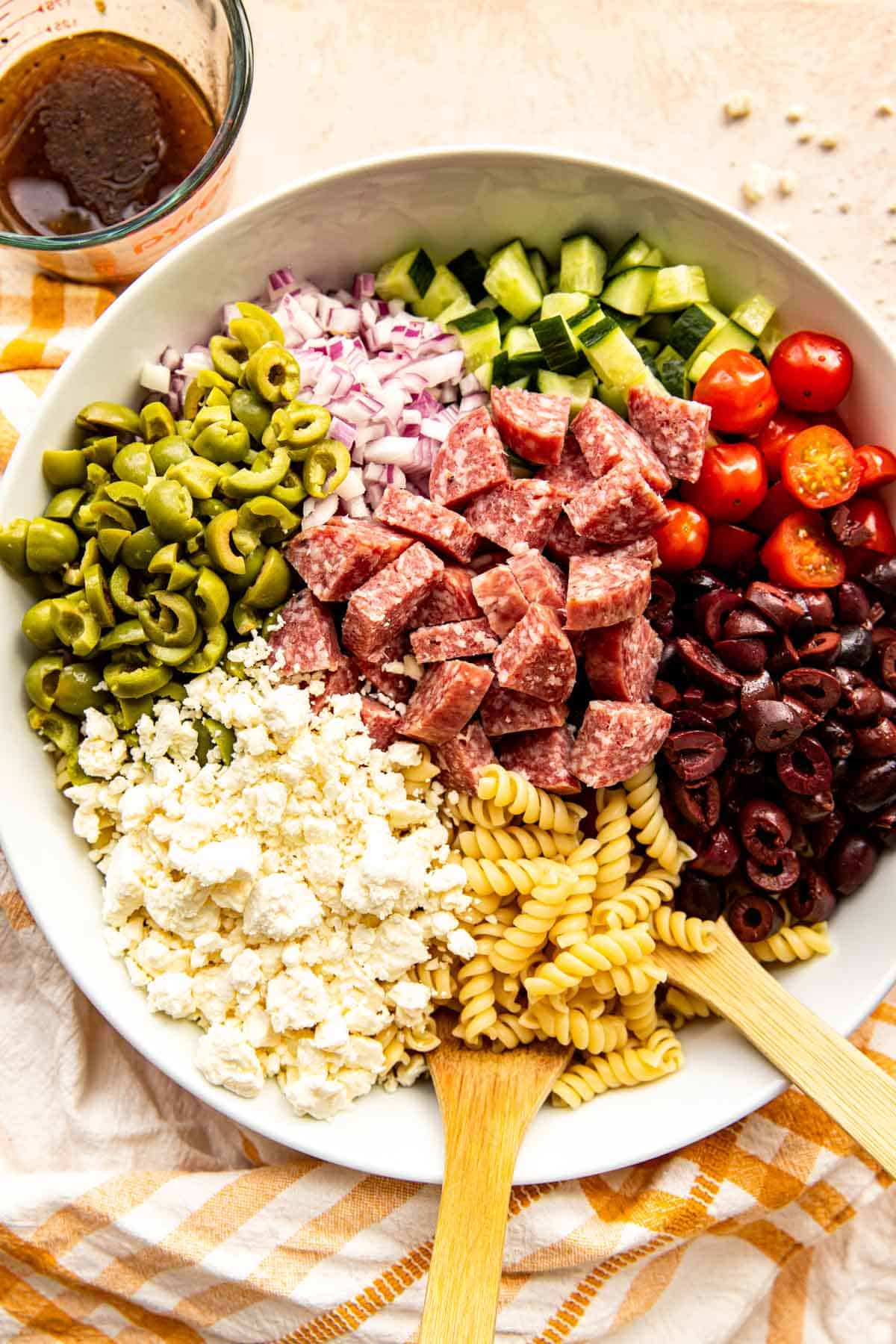 olives, red onions, tomatoes, salami, feta, and pasta in a large bowl with wooden spoons next to a measuring cup with dressing