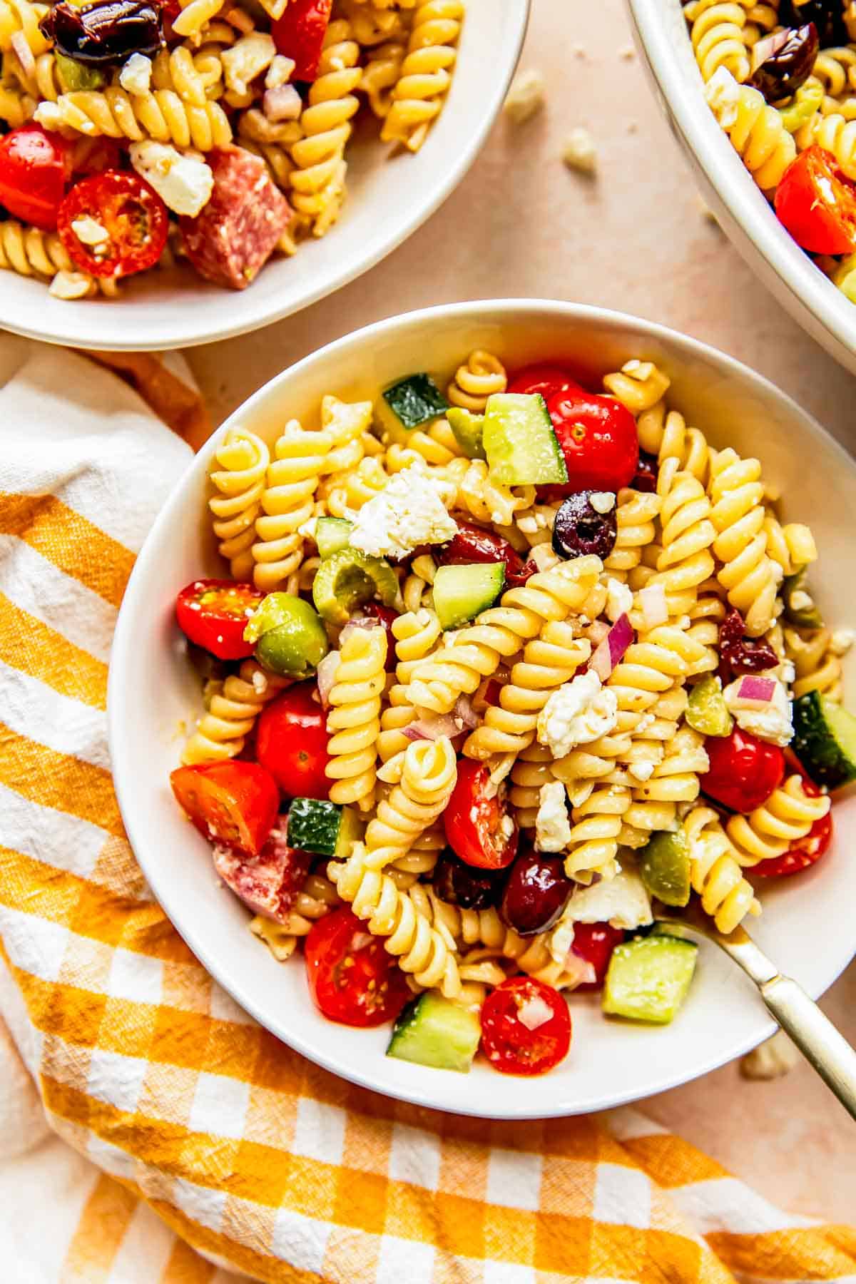 mediterranean pasta salad in a white bowl next to a gingham yellow towel