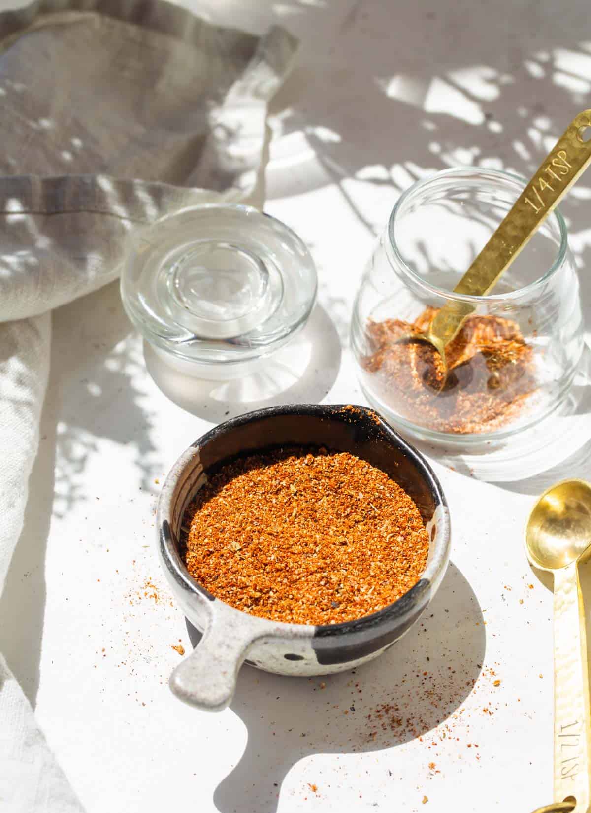 taco seasoning in a black and grey measuring cup next to a glass jar with a gold measuring spoon inside and additional taco seasoning