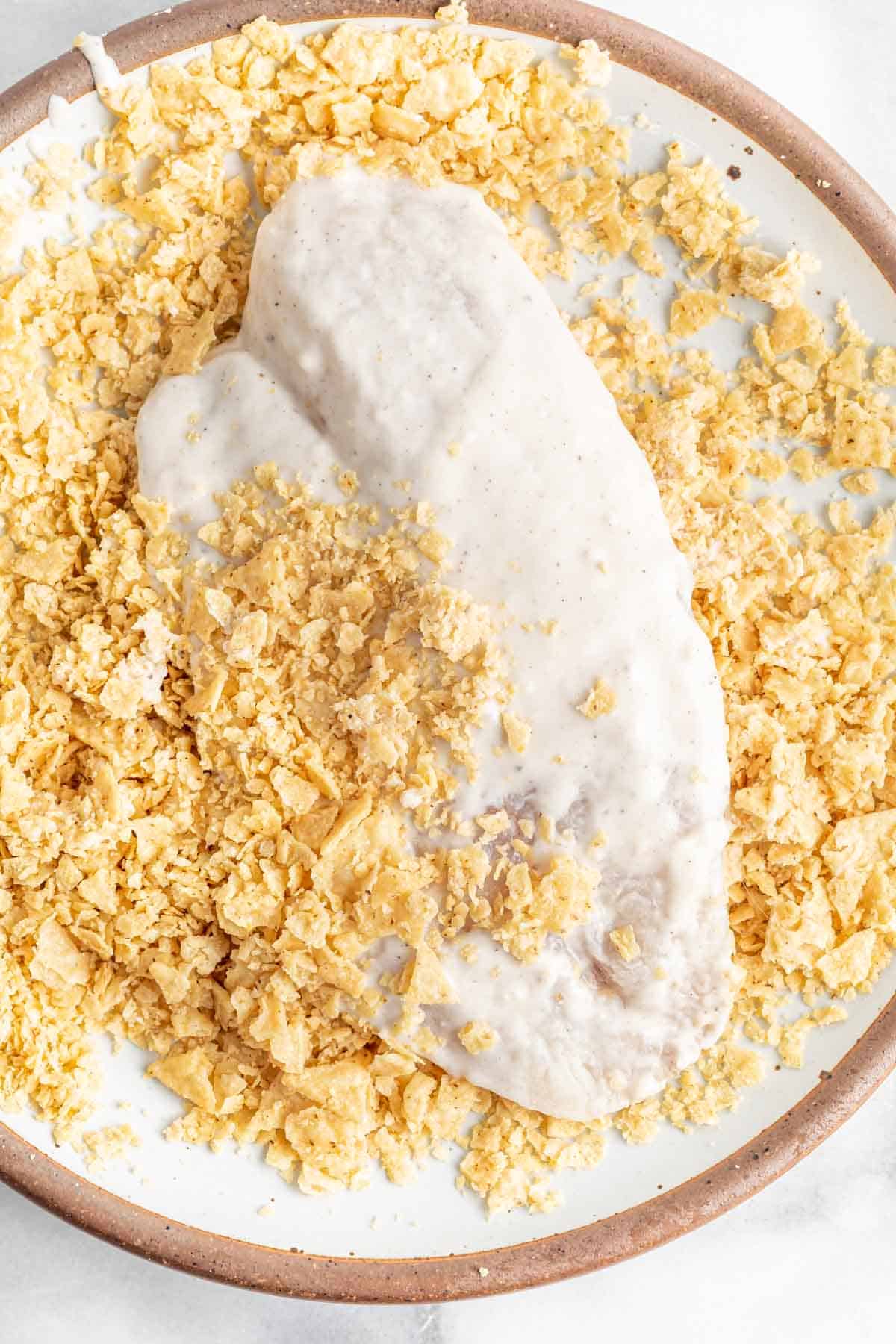 tilapia coated in batter being dipped into a plate of crushed tortilla chips