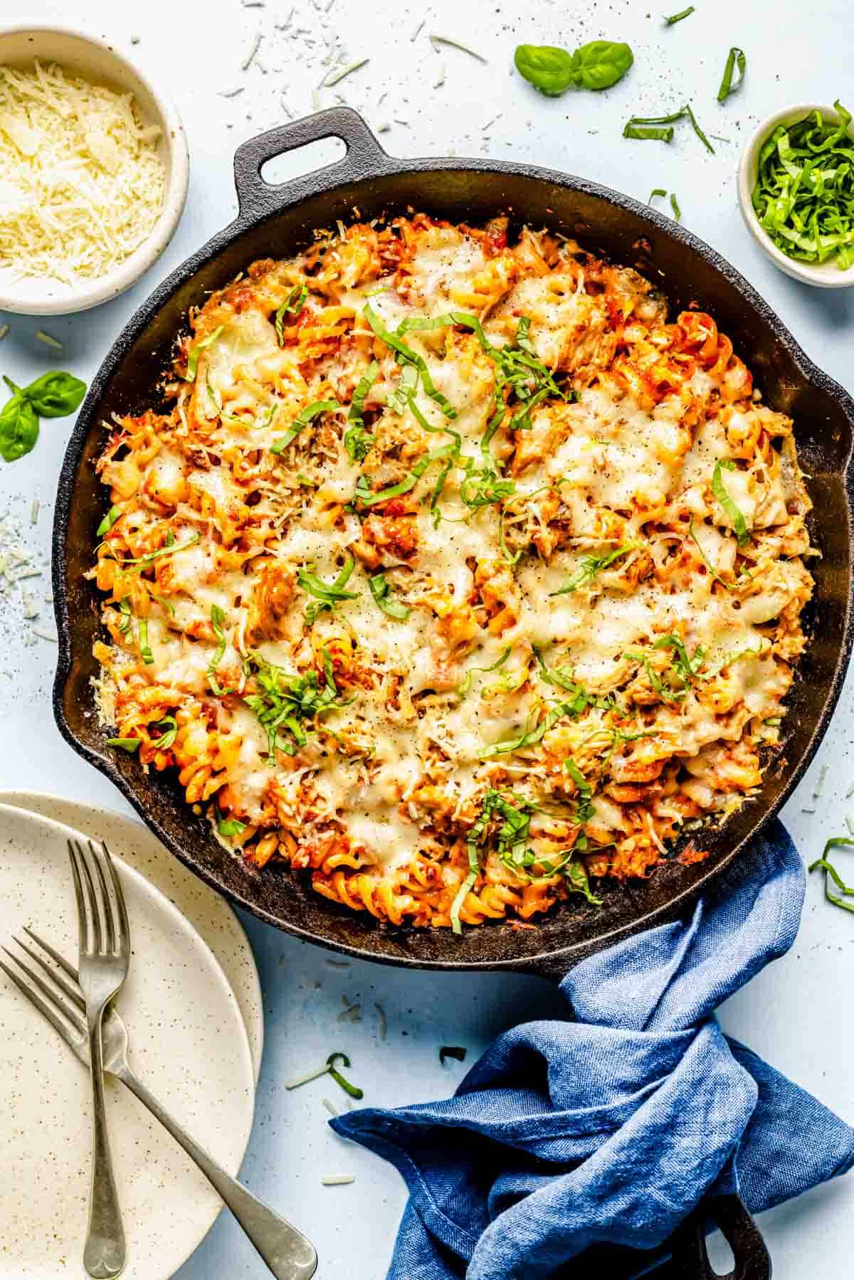 tuna pasta bake in a cast iron skillet topped with fresh basil next to a bowl of parmesan cheese and plates