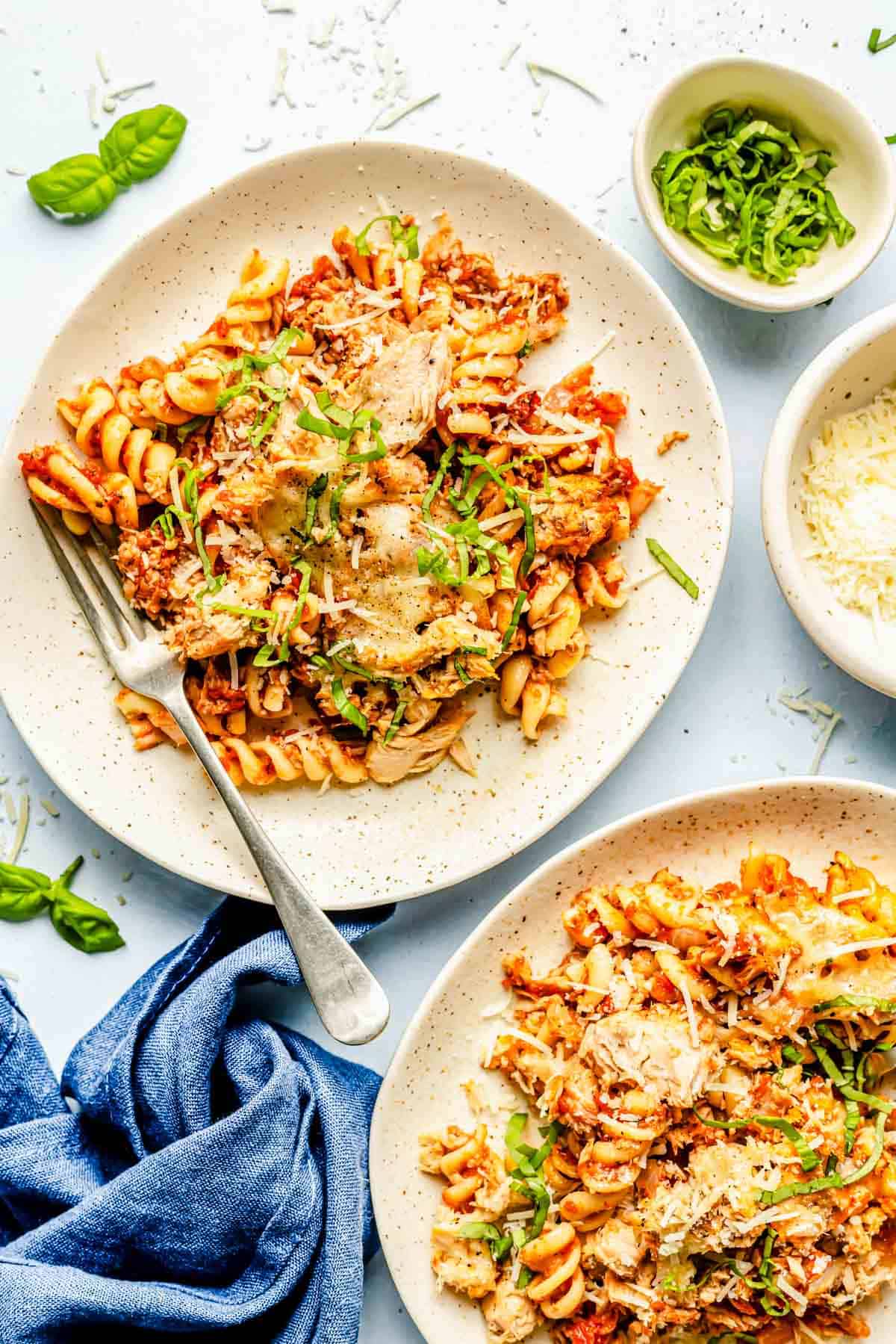 tuna pasta bake plated on two plates with a metal fork next to bowls of fresh basil and parmesan cheese