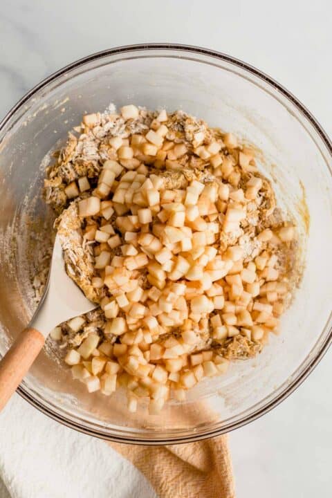 diced apples are being folded into the oats and cookie dough in a clear bowl