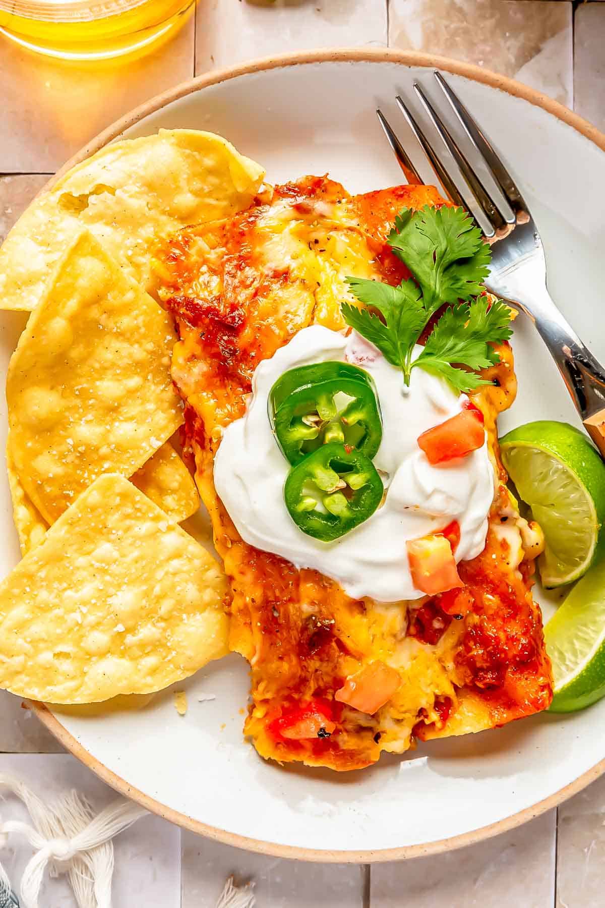 two beef enchiladas on a white plate topped with sour cream, jalapenos, and tomatoes next to corn tortilla chips and fresh lime slices