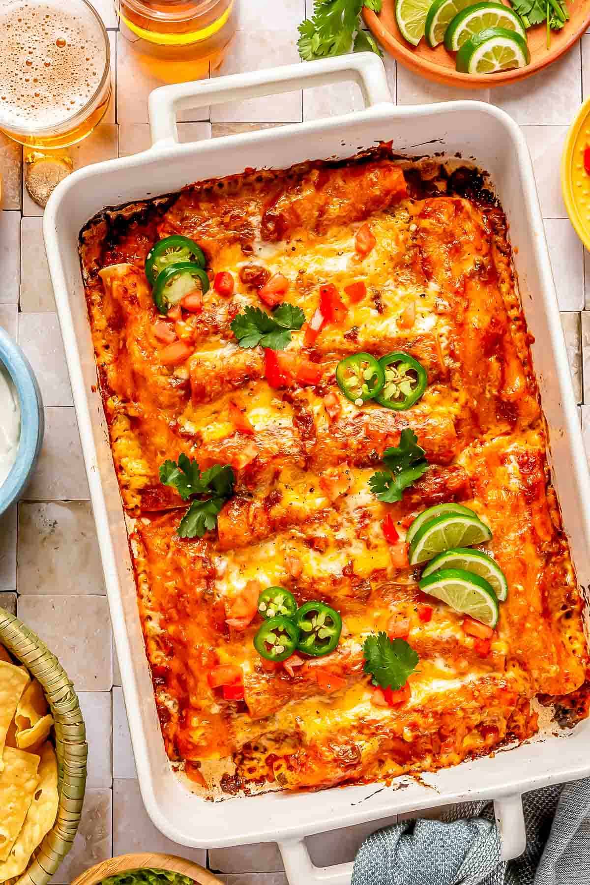 beef enchiladas in a baking dish topped with cilantro, jalapeños and lime slices next to an alcoholic beverage