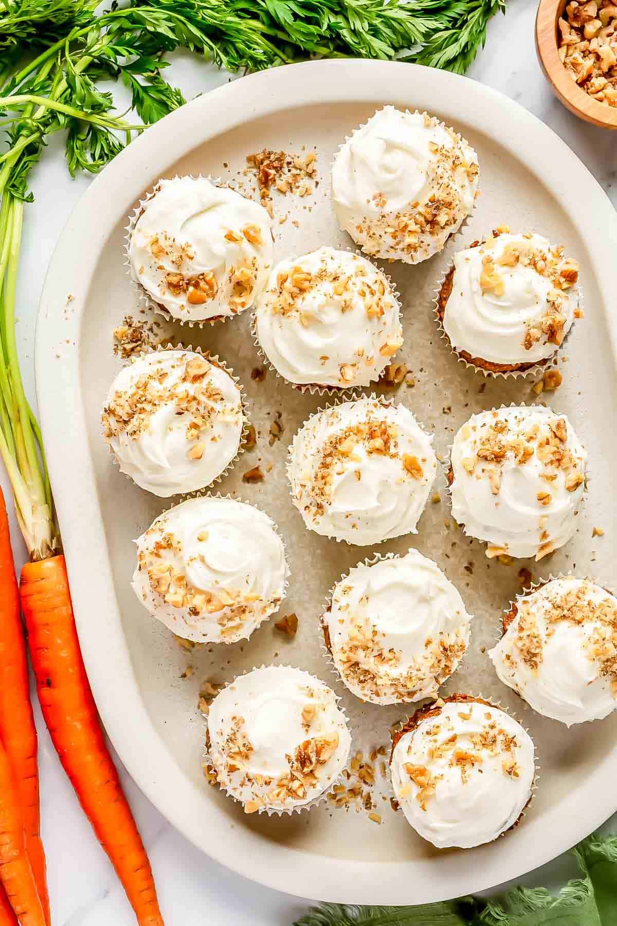 12 carrot cake muffins with cream cheese frosting topped with chopped walnuts on a large oval platter next to a large carrot