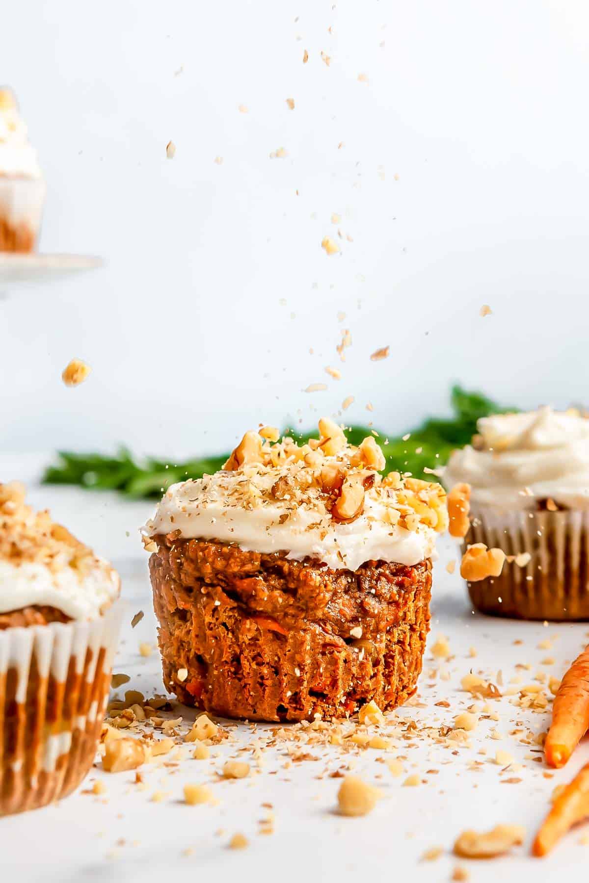 chopped walnuts being sprinkled on top of carrot cake muffins and cream cheese frosting