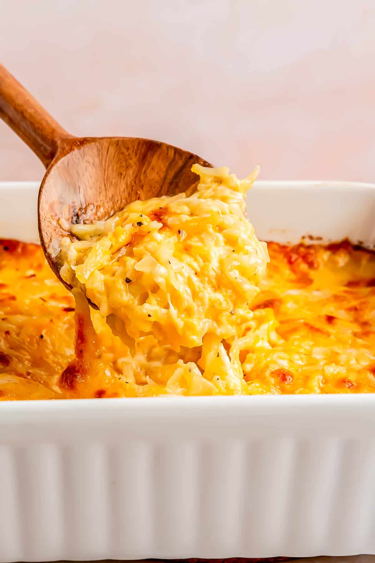 a wooden spoon scoops out cheesy hashbrown casserole from the baking dish