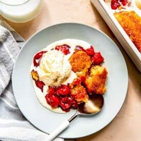 cherry cobbler on a blue plate with 1 scoop of melted vanilla ice cream and a metal spoon