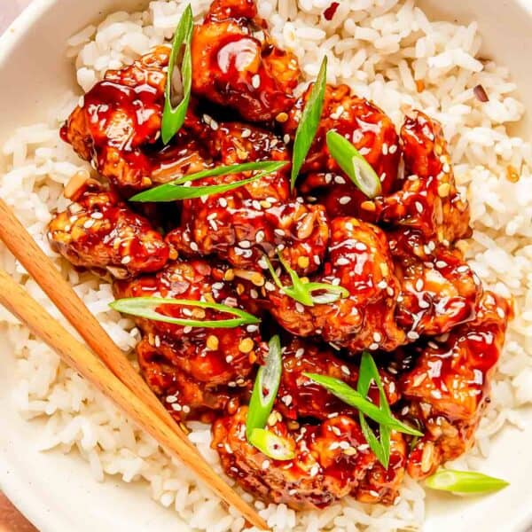 general tso's chicken on top of white rice in a bowl with wooden chopsticks. garnished with sesame seeds and scallions