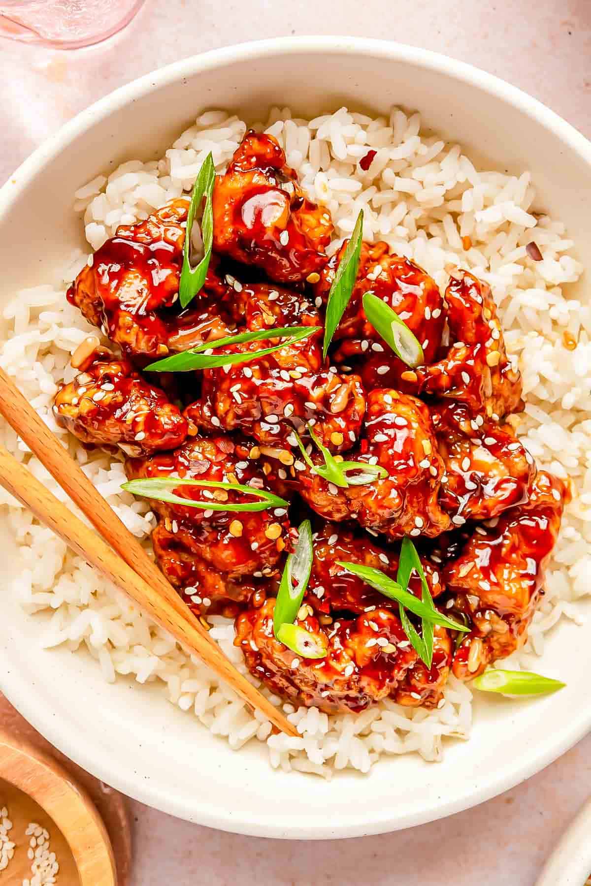 general tso's chicken on top of white rice in a bowl with wooden chopsticks. garnished with sesame seeds and scallions