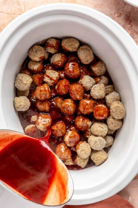 frozen meatballs in an insert of a slow cooker with grape jelly and chili sauce being poured on top