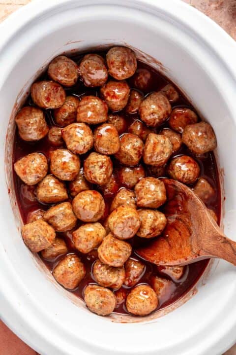 grape jelly and chili sauce stirred and coating frozen meatballs in a slow cooker insert with a wooden spoon