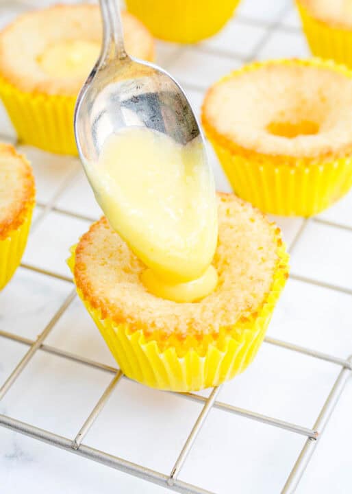 a metal spoonful of lemon curd is being dropped into a hole in the center of the cupcake
