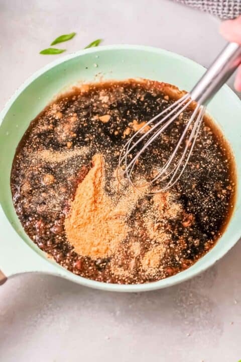 soy sauce, brown sugar, and other liquids in a green saucepan being whisked with a metal whisk