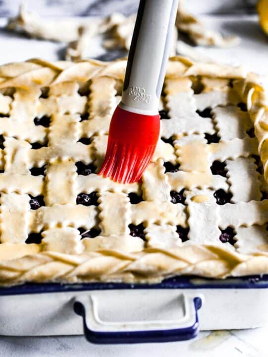 a red silicone pastry brush is brushing the top of the lattice design with egg wash