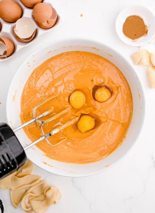 a hand mixer is in a bowl with orange batter and 3 eggs