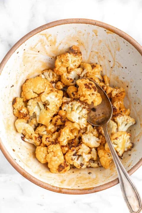 roasted cauliflower being tossed in buffalo sauce in a ceramic bowl with a metal spoon
