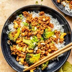 cashew chicken and broccoli in a black bowl with white rice next to wooden chopsticks
