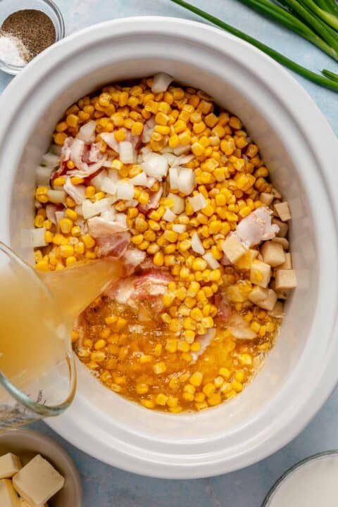 corn kernels, onion, bacon, and potatoes in the insert of a slow cooker with chicken broth being poured in