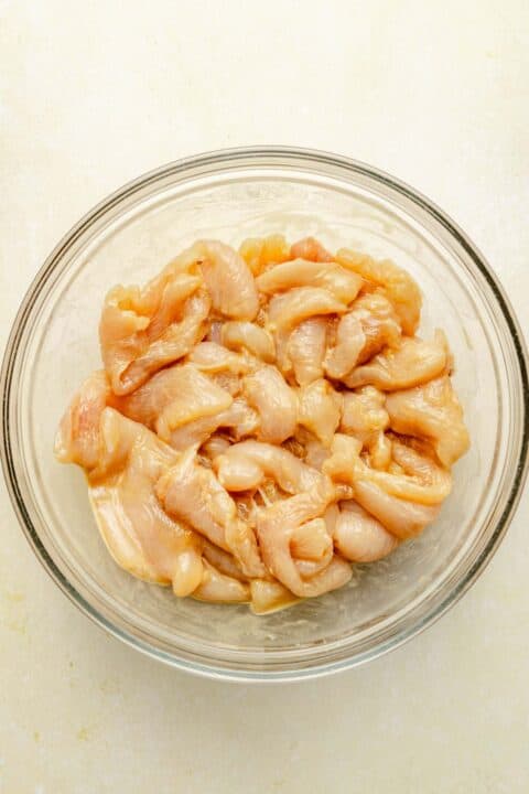 raw thin slices of chicken marinating in a clear bowl