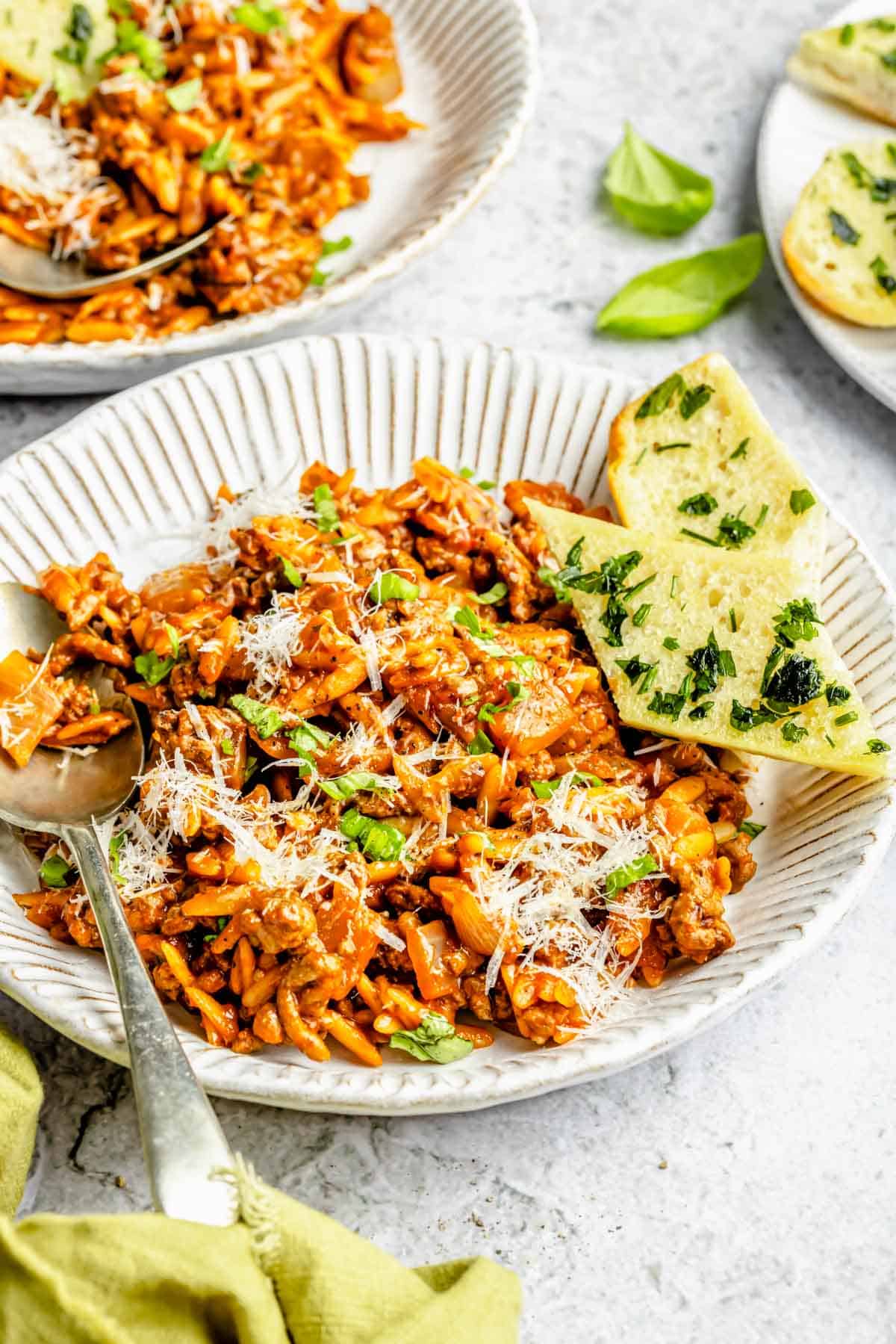 ground beef and orzo in a white plate next to a metal spoon and garlic bread next to the pasta