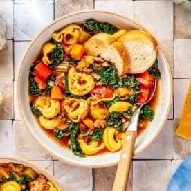 a bowl with a metal spoon has sausage tortellini soup and sliced bread on the inside