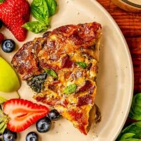 a slice of bacon mushroom spinach quiche on a beige plate next to fresh strawberries, blueberries, and apples.