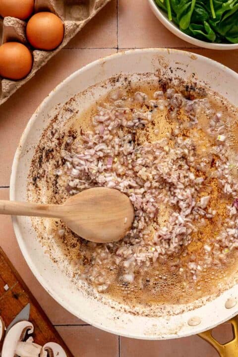 shallots being cooked in bacon grease in a white skillet with a wooden spoon