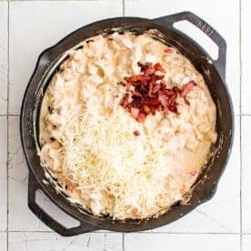 shredded mozzarella cheese and bacon pieces on top of cooked chicken mixture in cast iron skillet