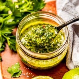a black spoon has a scoop of chimichurri sauce scooped up from inside a glass jar