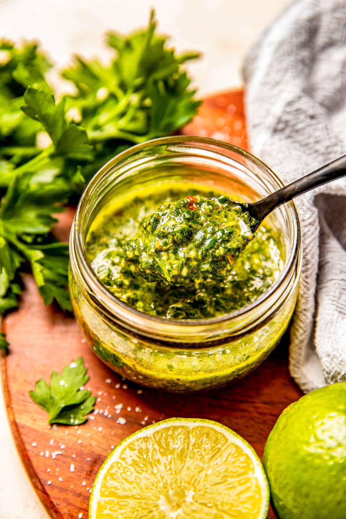 a black spoon has a scoop of chimichurri sauce scooped up from inside a glass jar