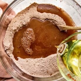 vegetable oil is being poured into cocoa powder and flour mixture in a clear bowl with a wooden spoon