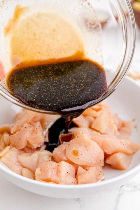 soy sauce mixture is being poured on cubed raw chicken in a white bowl