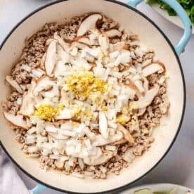 garlic, onion, ginger, and mushrooms are added on top of the ground pork in an enameled cast iron skillet