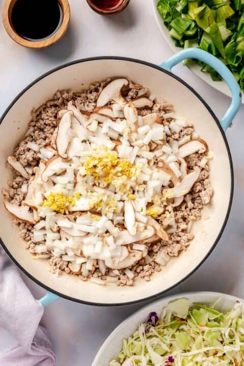 garlic, onion, ginger, and mushrooms are added on top of the ground pork in an enameled cast iron skillet