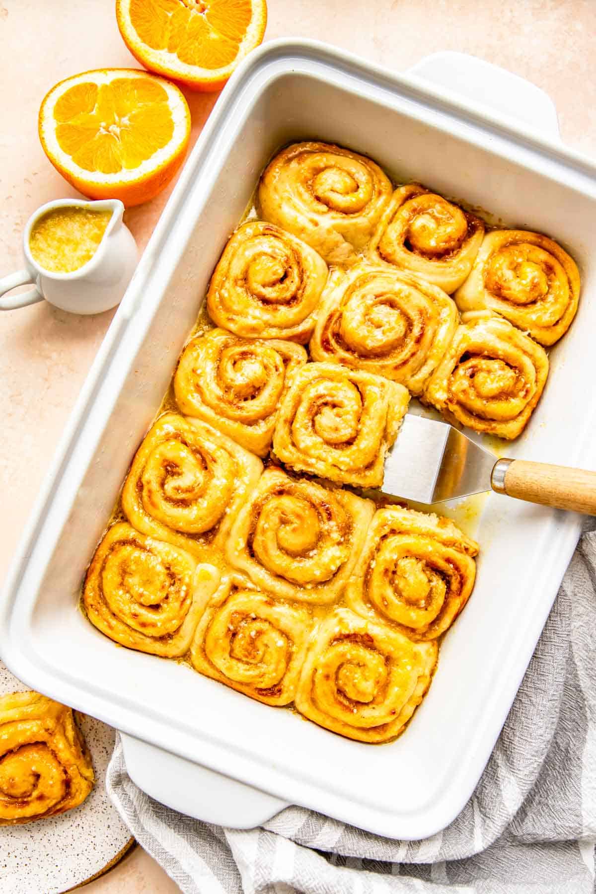 orange rolls baked and glazed in a white baking dish with a metal spatula holding one