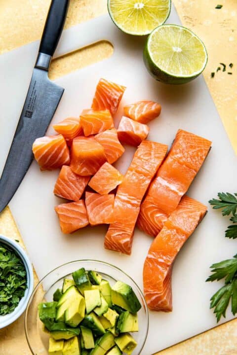 fresh salmon filets and salmon cubes on a cutting board next to a knife and limes and avocados
