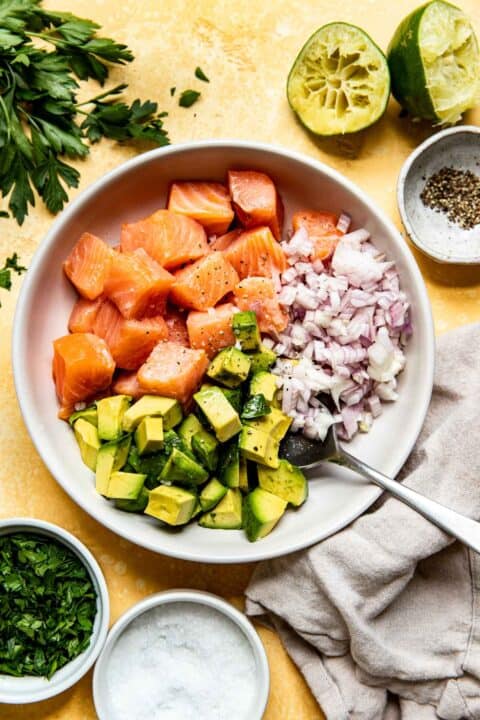 cubed salmon, avocado, shallots, and metal spoon in a bowl next to freshly juiced limes and a linen towel