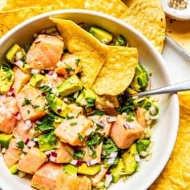 an overhead image of salmon ceviche in a white bowl with a metal spoon and two tortilla chips in the bowl