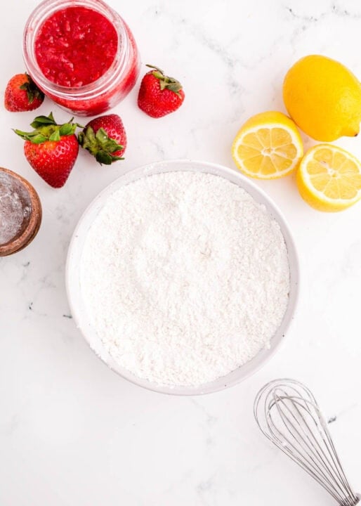 dry ingredients in a white bowl next to a metal whisk and fresh strawberries and lemons