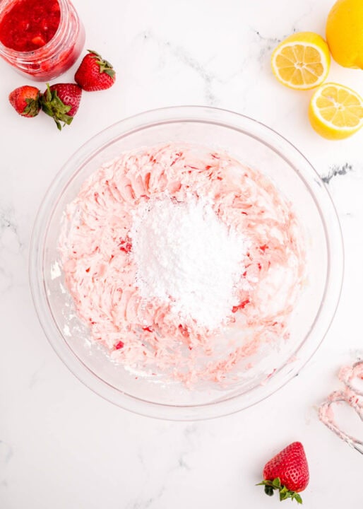additional powdered sugar is added to the pink strawberry frosting mixture in a clear bowl