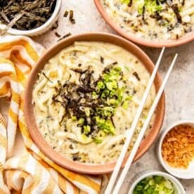 udon noodle soup in a clay bowl topped with seaweed and green onions with wooden chopsticks sitting on the bowl