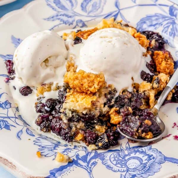 two scoops of melted vanilla ice cream on top of blueberry dump cake on a blue china plate with a metal spoon