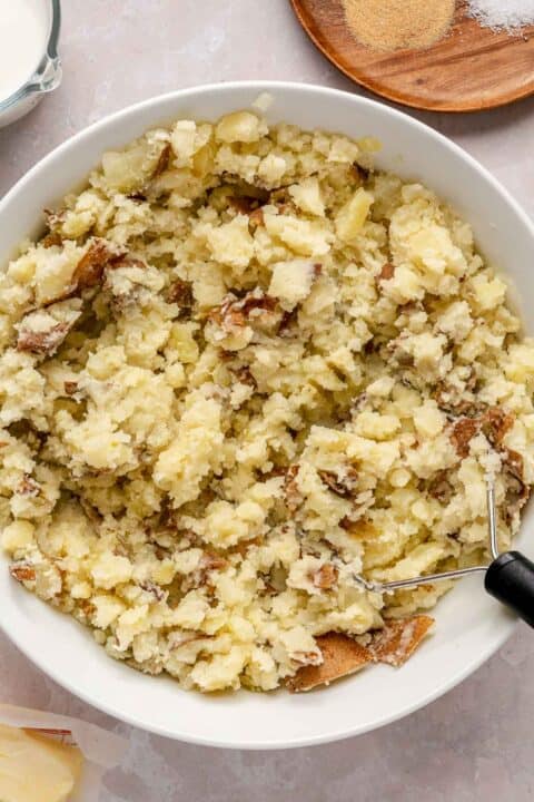 boiled potatoes mashed in a white bowl with a potato masher