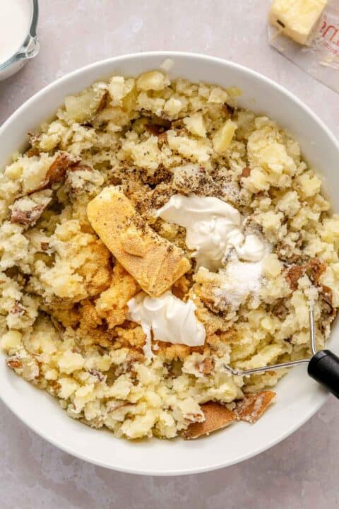 butter, sour cream, and seasonings are added to the potatoes in the white bowl