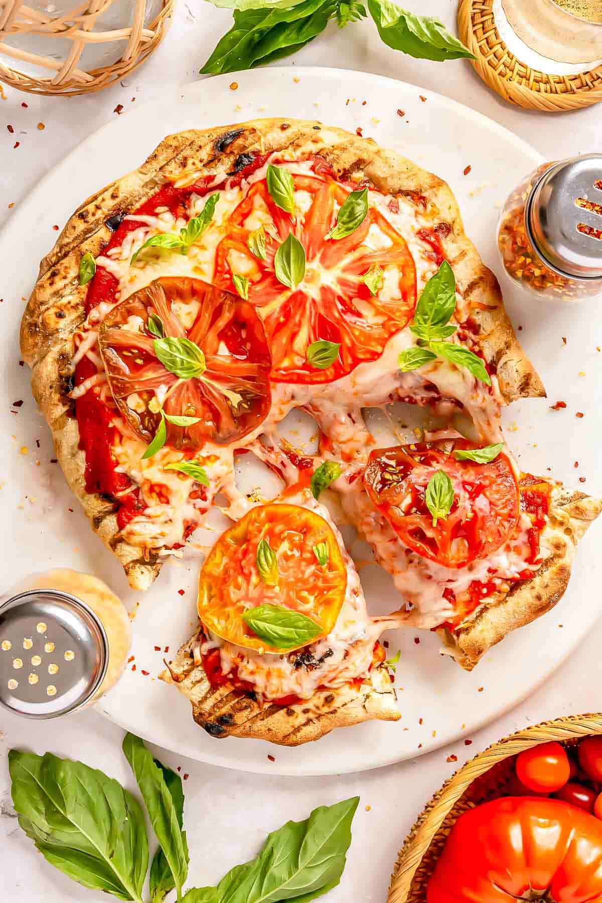 grilled caprese pizza topped with fresh basil leaves sliced into wedges on a large white plate