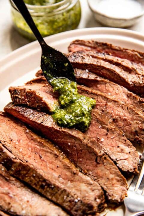 a black spoon spoons chimichurri sauce on top of sliced flank steak pieces