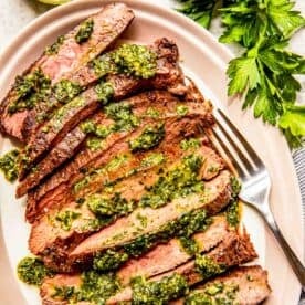 sliced flank steak strips are arranged on a pink plate with chimichurri sauce on top. the plate has a metal fork on it